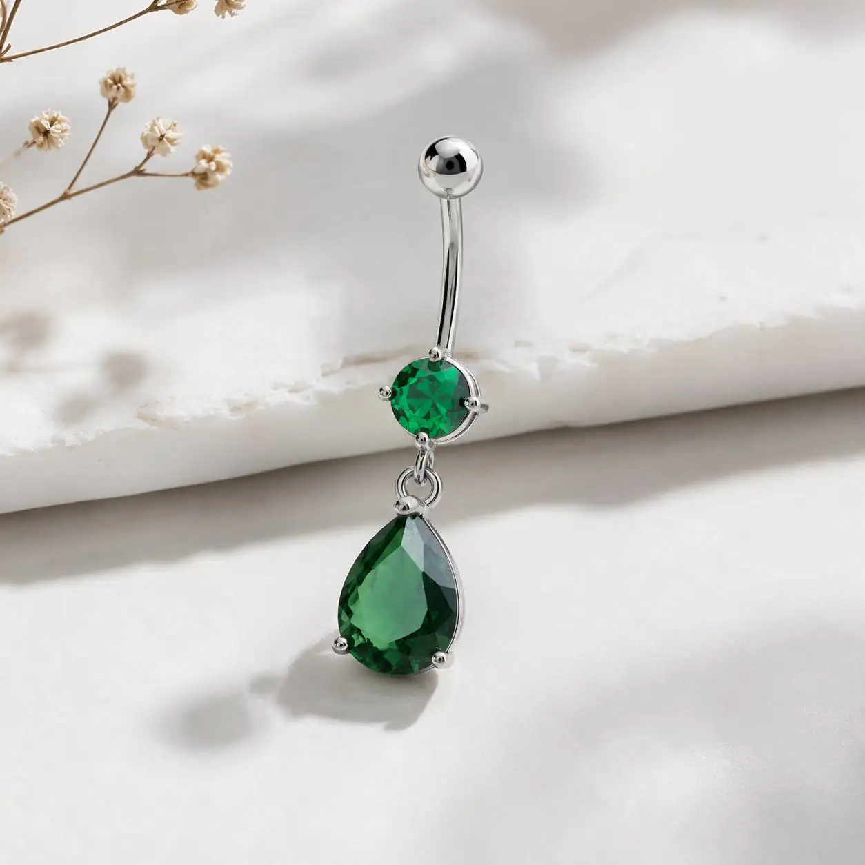 emerald-green-dangle-navel-piercing-closeup نمای نزدیک پیرسینگ ناف نگین سبز با آویز اشکی و میله منحنی نقرهای