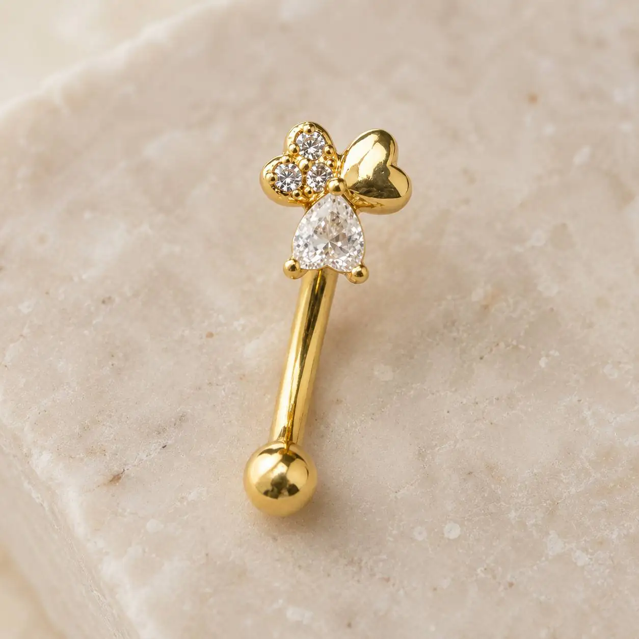 gold-eyebrow-piercing-on-marble نمای نزدیک پیرسینگ ابرو طلایی با نگینهای شفاف روی سنگ روشن