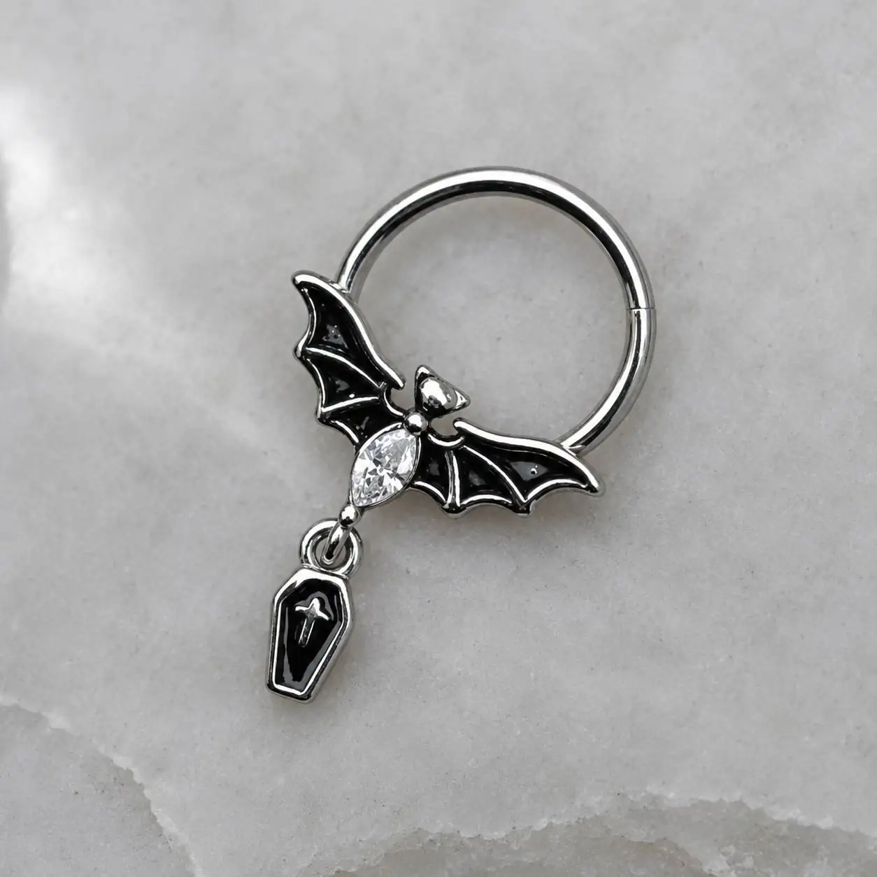 silver-bat-coffin-septum-jewelry-stone پیرسینگ سپتوم گاتیک با بال خفاش مشکی، نگین شفاف و آویز تابوت روی سطح روشن سنگی
