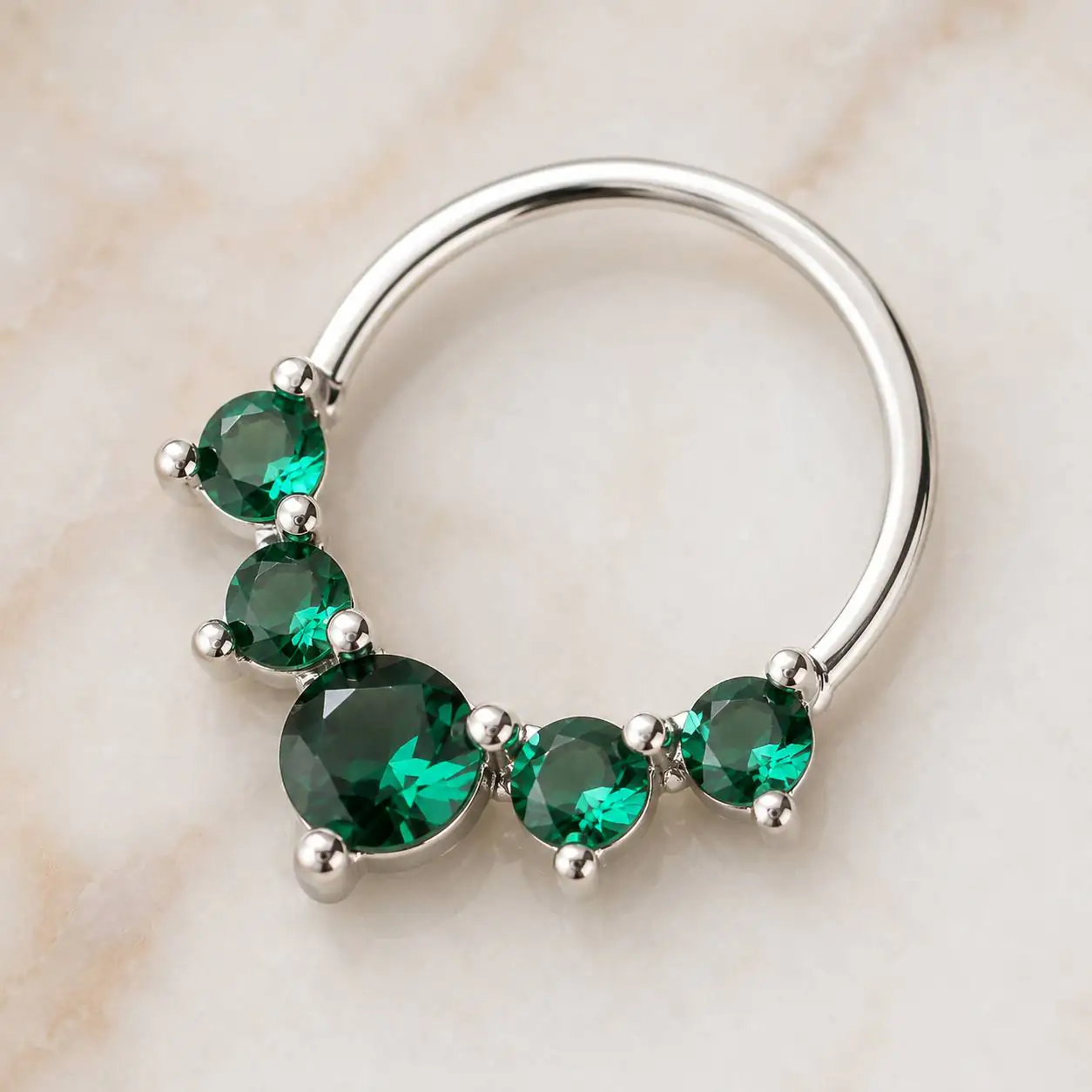 silver-emerald-septum-ring-product-photo پیرسینگ سپتوم نقرهای با نگینهای سبز زمردی روی پسزمینه روشن