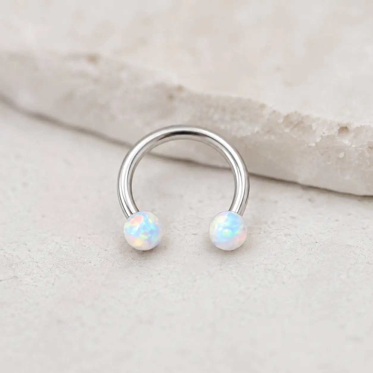 silver-opal-smiley-piercing-white-stone پیرسینگ اسمایل حلقهای نقرهای با دو مهره اپال سفید روی زمینه سنگی روشن